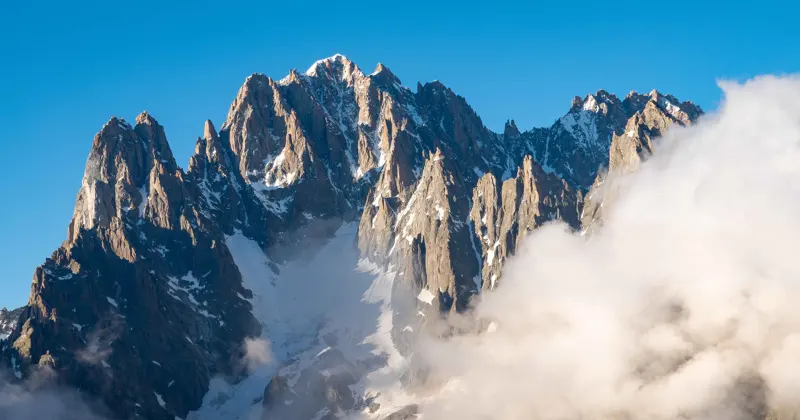 Gekartelde, met sneeuw bestrooide bergtoppen steken scherp af tegen een helderblauwe hemel, terwijl witte wolken de lagere hellingen gedeeltelijk aan het zicht onttrekken.